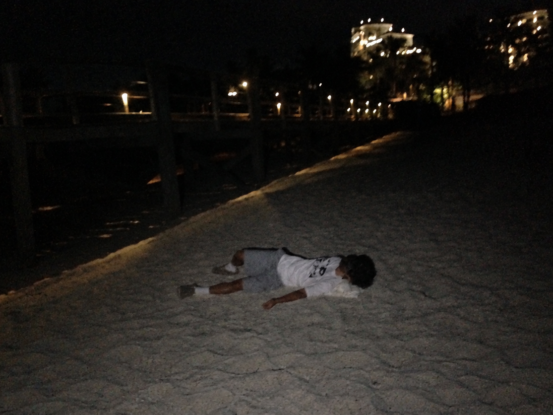 A person lying on their back on a sandy beach at night, wearing a white t-shirt and gray shorts. The background is dark with beachfront buildings and lights visible in the distance.